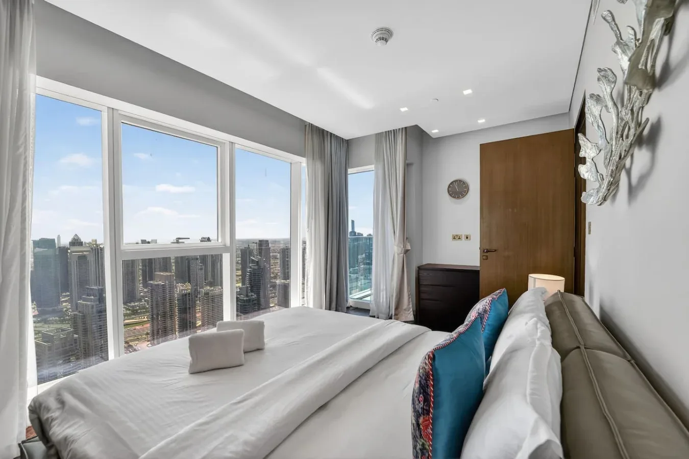 A bright bedroom with a bed beside large windows overlooking the city.