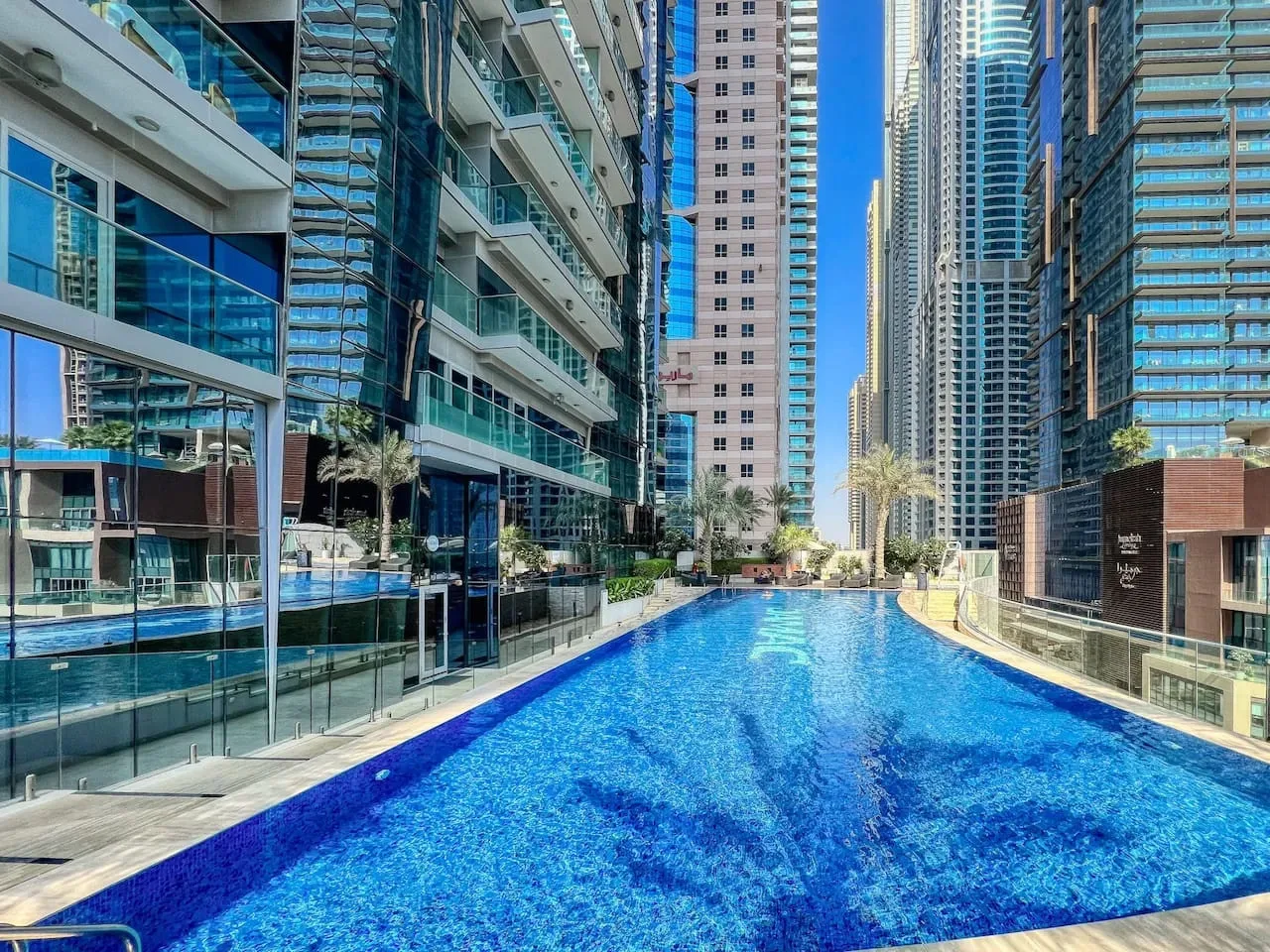 A swimming pool sits among tall modern buildings, with glass-fronted facades surrounding the water.