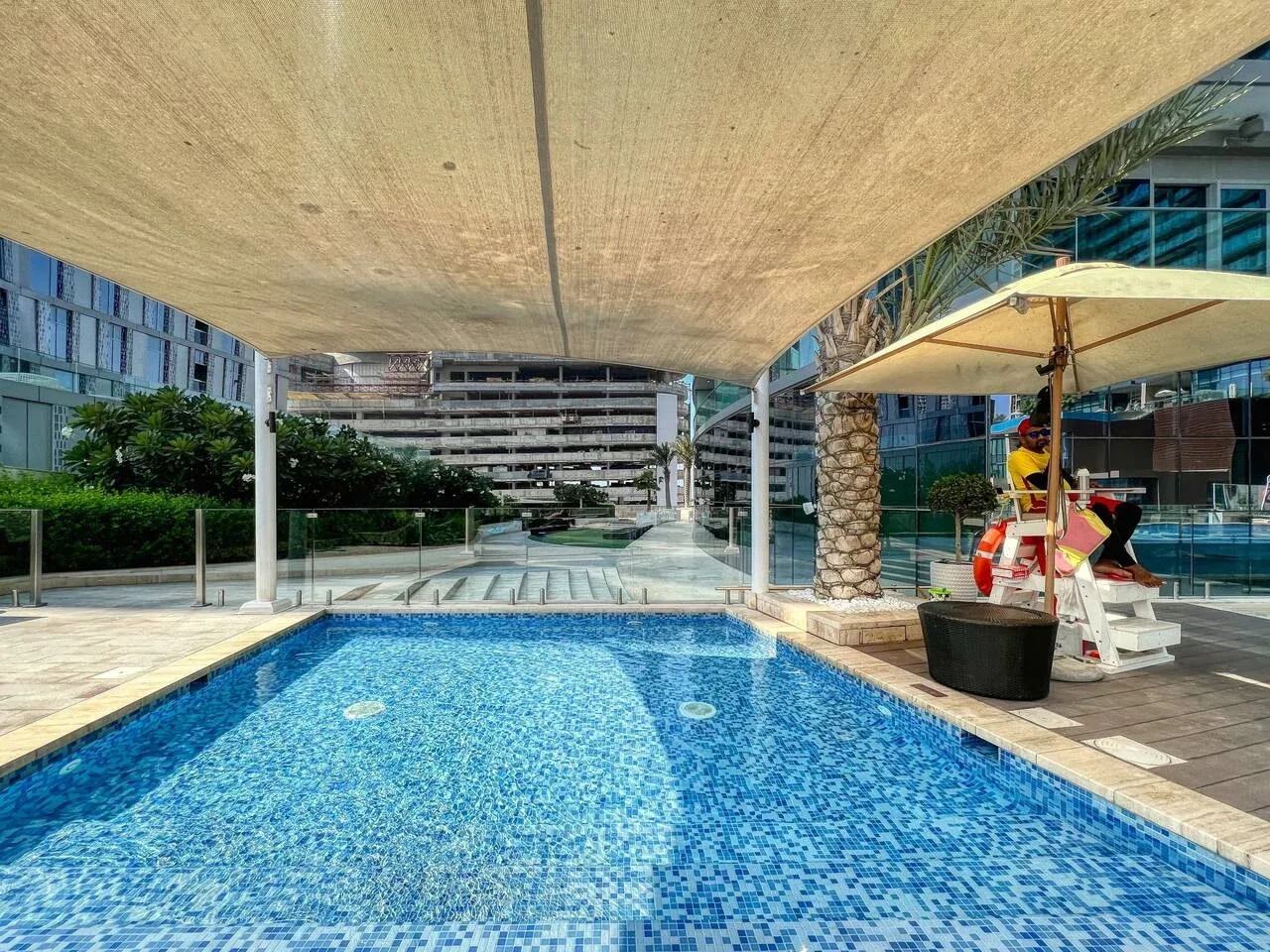 An outdoor swimming pool sits beneath a broad shade canopy, with modern buildings nearby.