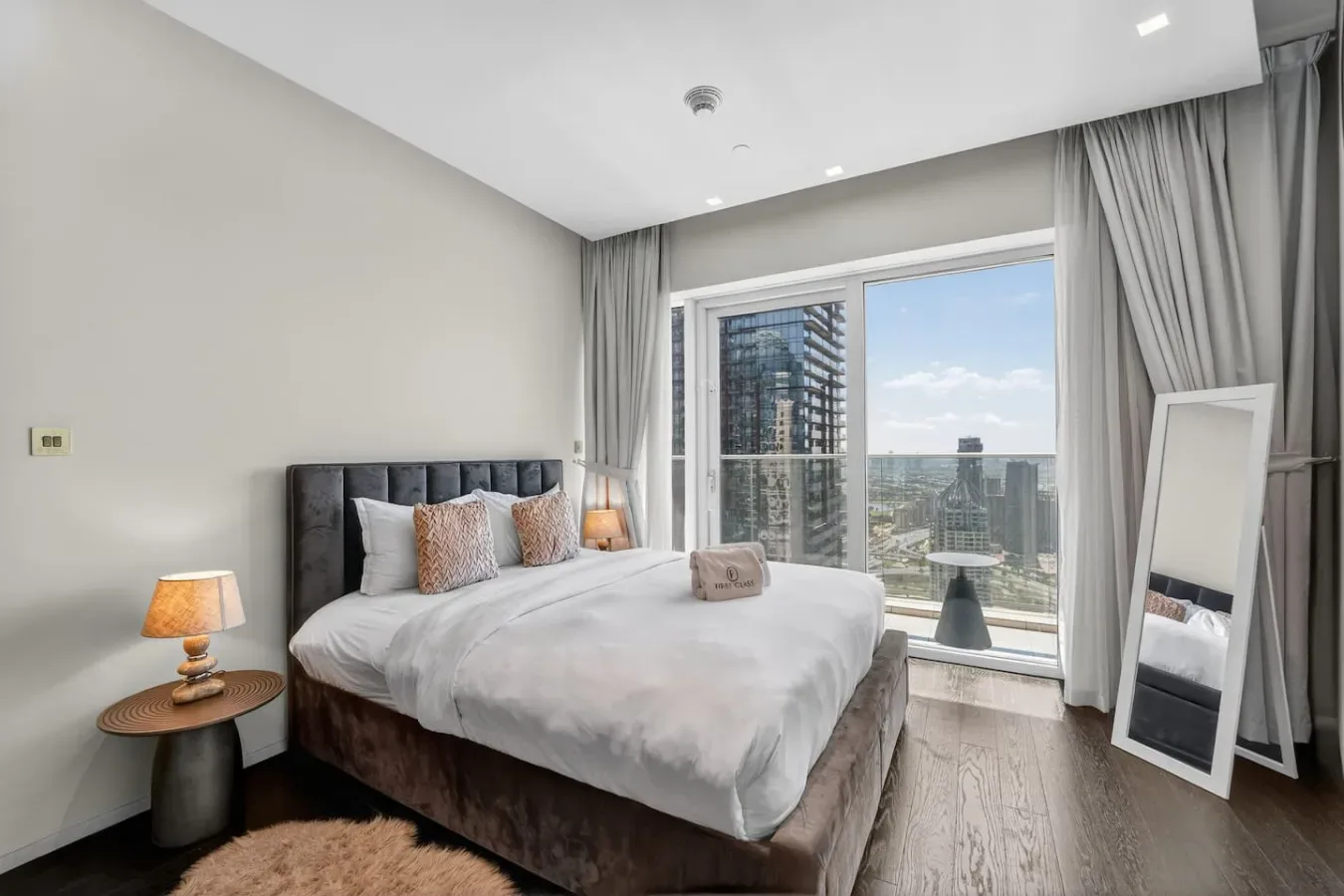 A bedroom with a bed beside large windows overlooking nearby buildings.