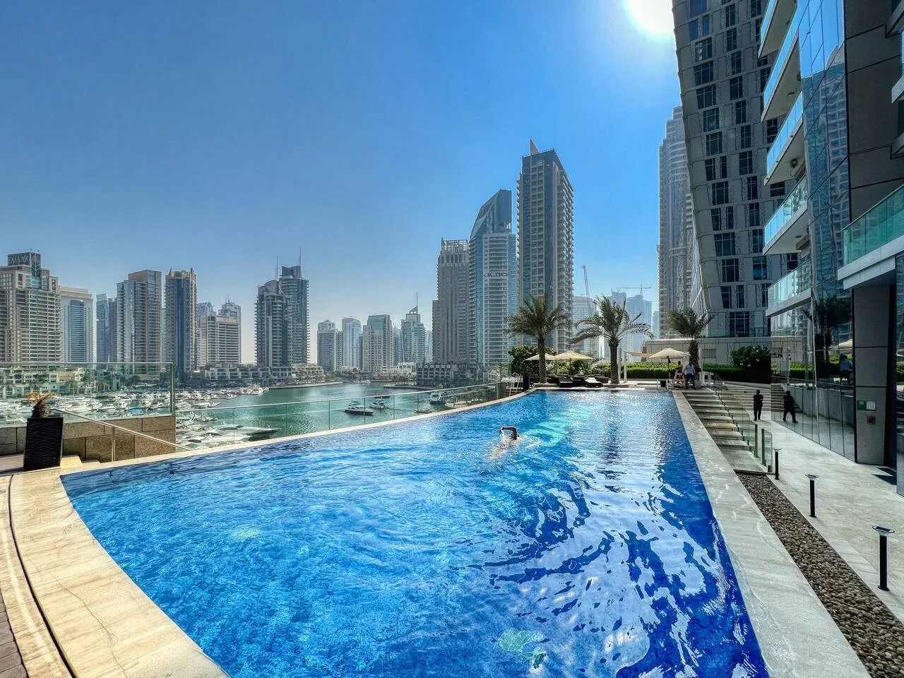 A rooftop swimming pool overlooks a waterfront skyline on a sunny day.