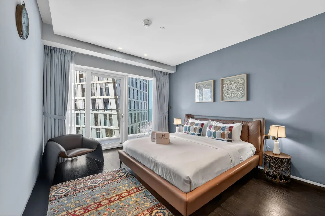 A bedroom with a large bed, colorful rug, and windows overlooking nearby buildings.