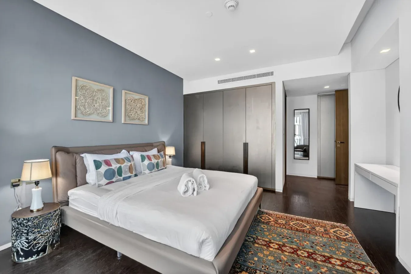 A bedroom with a neatly made bed, framed artwork, and a colorful rug.
