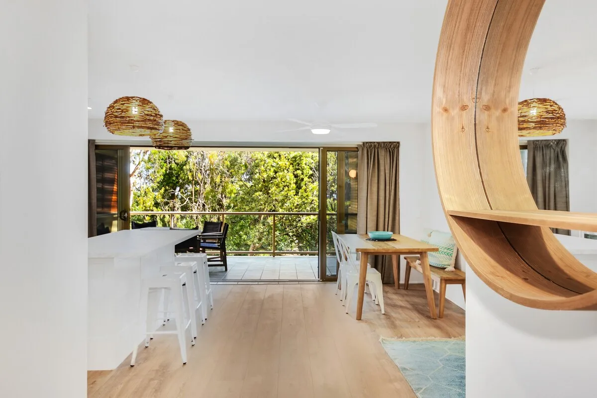 Bright open-plan living and dining area with a kitchen and windows overlooking greenery.