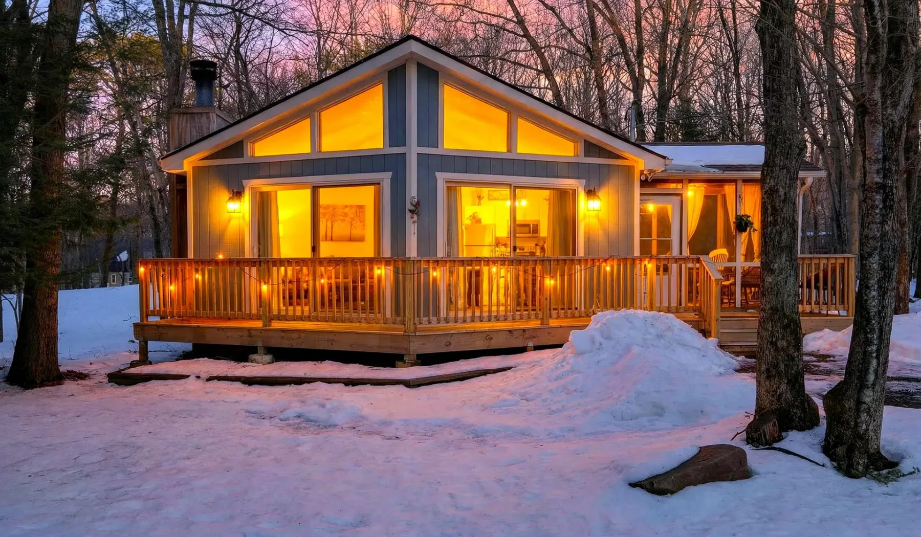 A warmly lit house with a wooden deck stands among snow-covered trees at dusk.