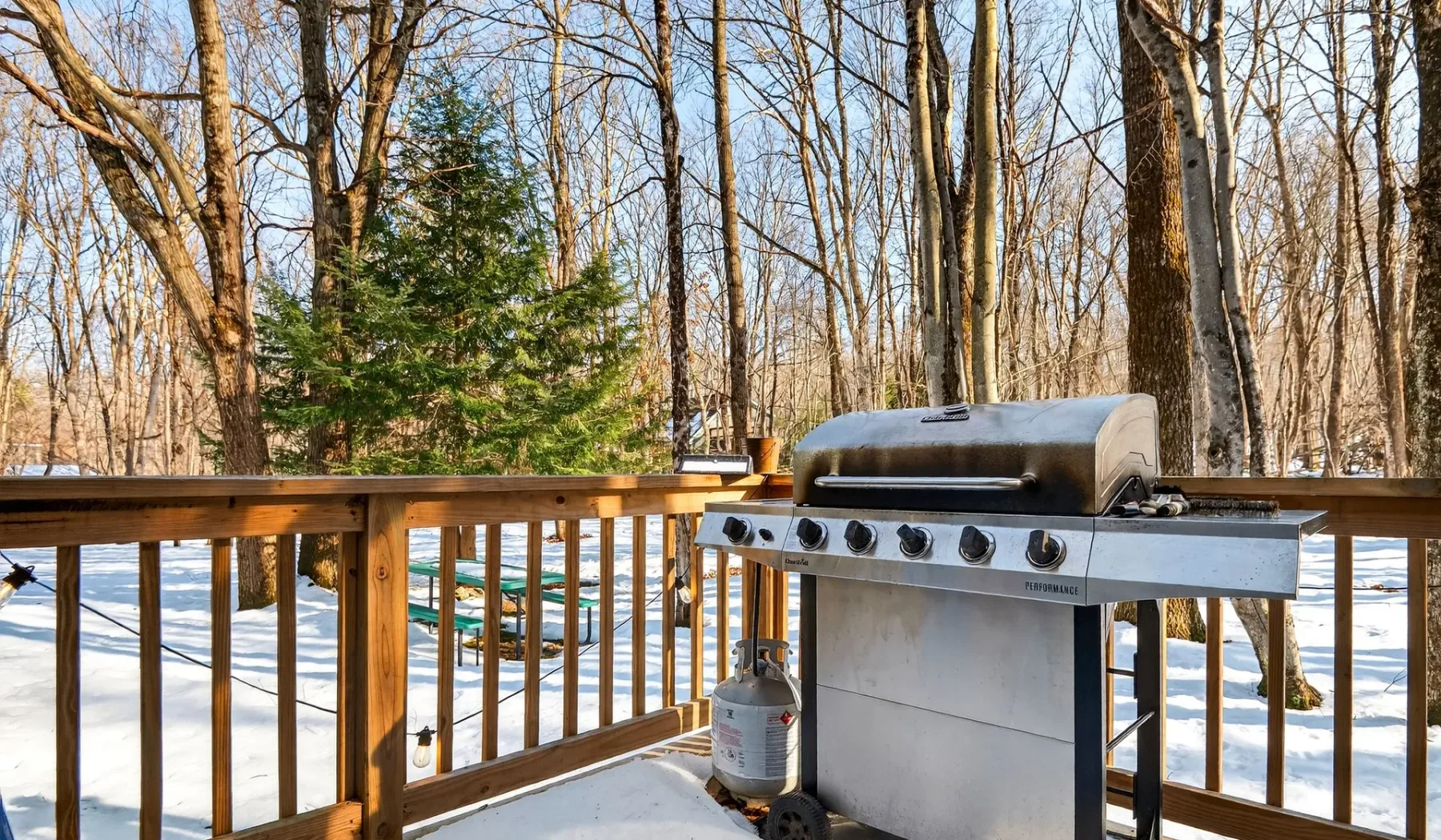 A barbecue grill sits on a wooden deck overlooking a snowy, wooded yard.