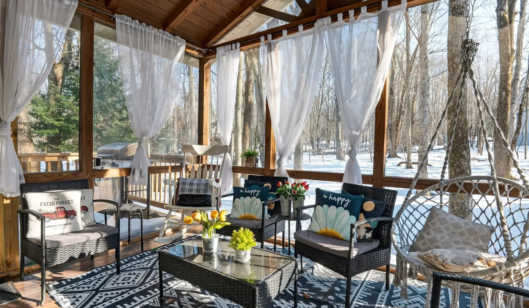 A sunroom with seating, sheer curtains, and views of a snowy wooded landscape.