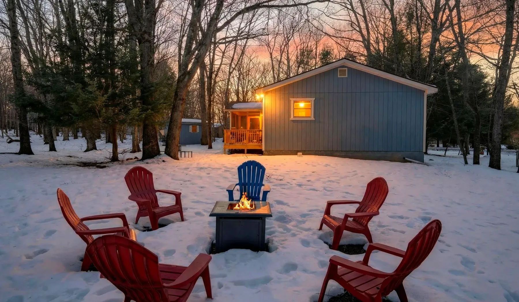 A snowy outdoor seating area with a small fire in front of a cabin at dusk.