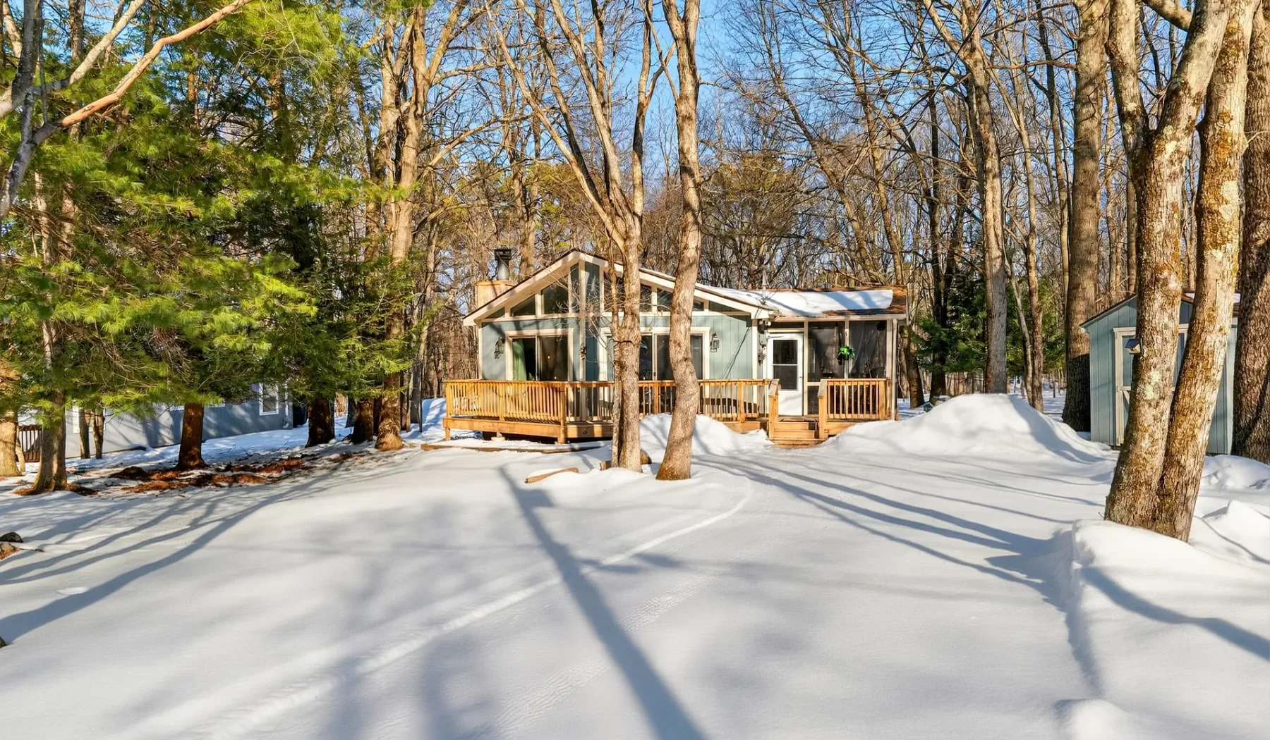 A house with a wooden deck sits among trees in a snow-covered yard.
