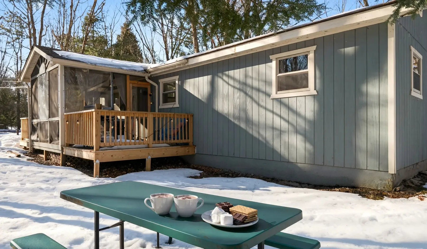A gray cabin with a wooden porch stands beside a snow-covered outdoor table.