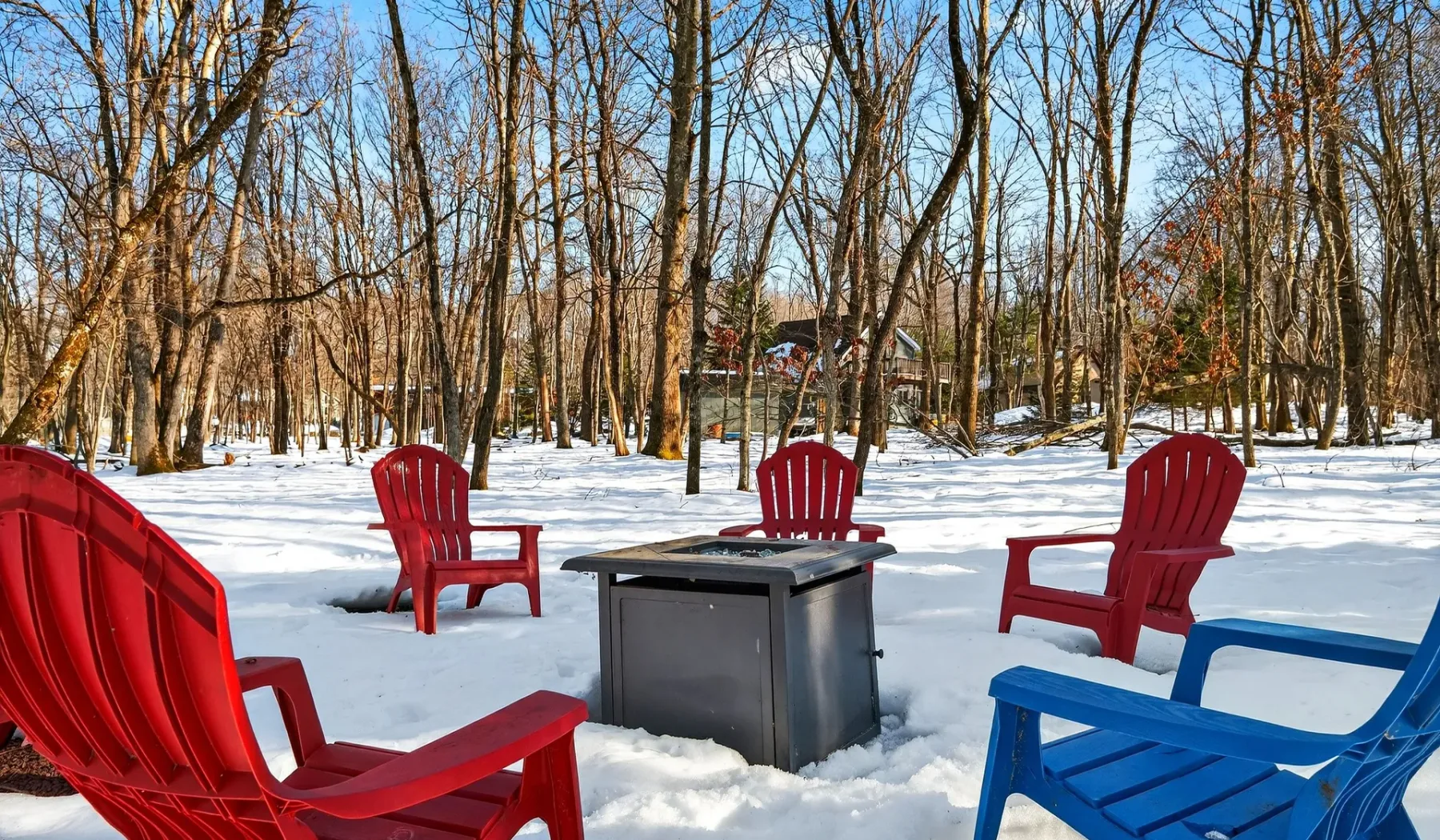 Snowy wooded outdoor seating area with colorful chairs around a fire pit.