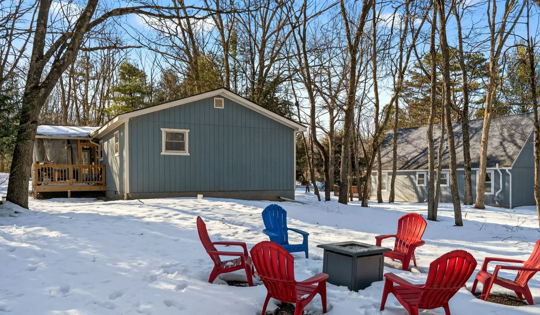 Snow-covered outdoor seating area between blue cabins in a leafless wooded setting.