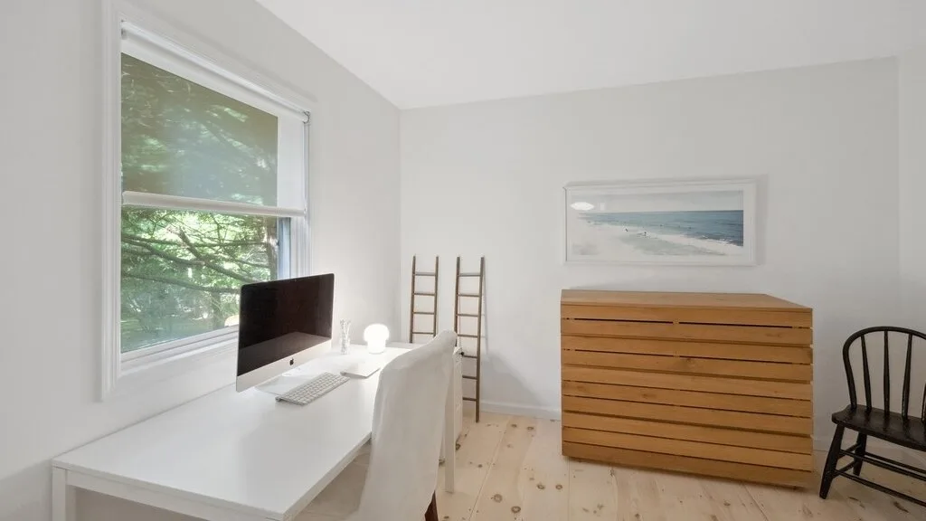 Un espacio de trabajo luminoso tiene un escritorio blanco junto a una ventana y una cómoda de madera.