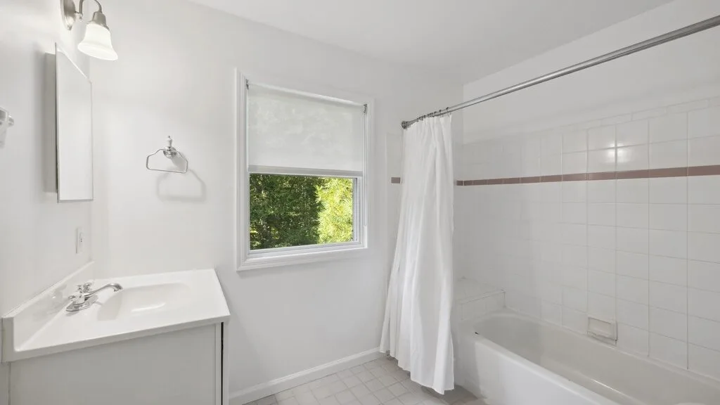 Un baño luminoso con lavabo, ventana y bañera detrás de una cortina de ducha.