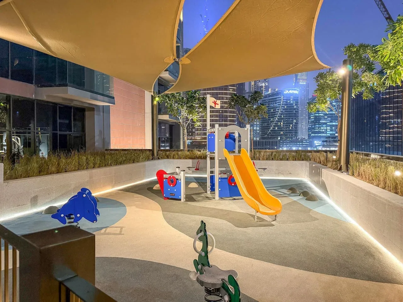 A rooftop playground with a slide and colorful play equipment beneath shade sails.
