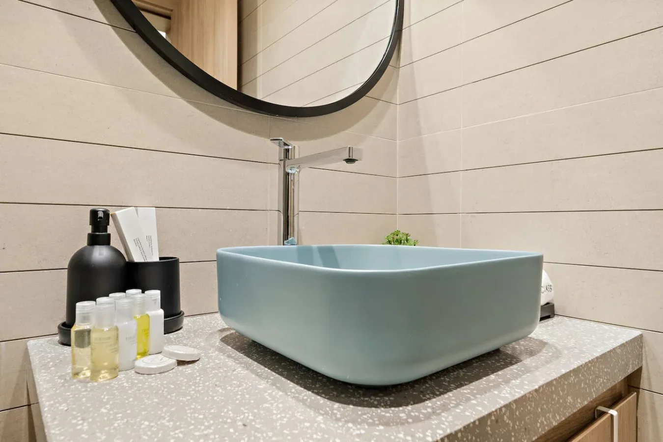 A bathroom vanity with a pale blue vessel sink, toiletries, and a round mirror.
