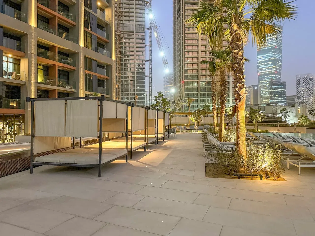 An outdoor terrace with lounge seating, palm trees, and illuminated high-rise buildings at dusk.