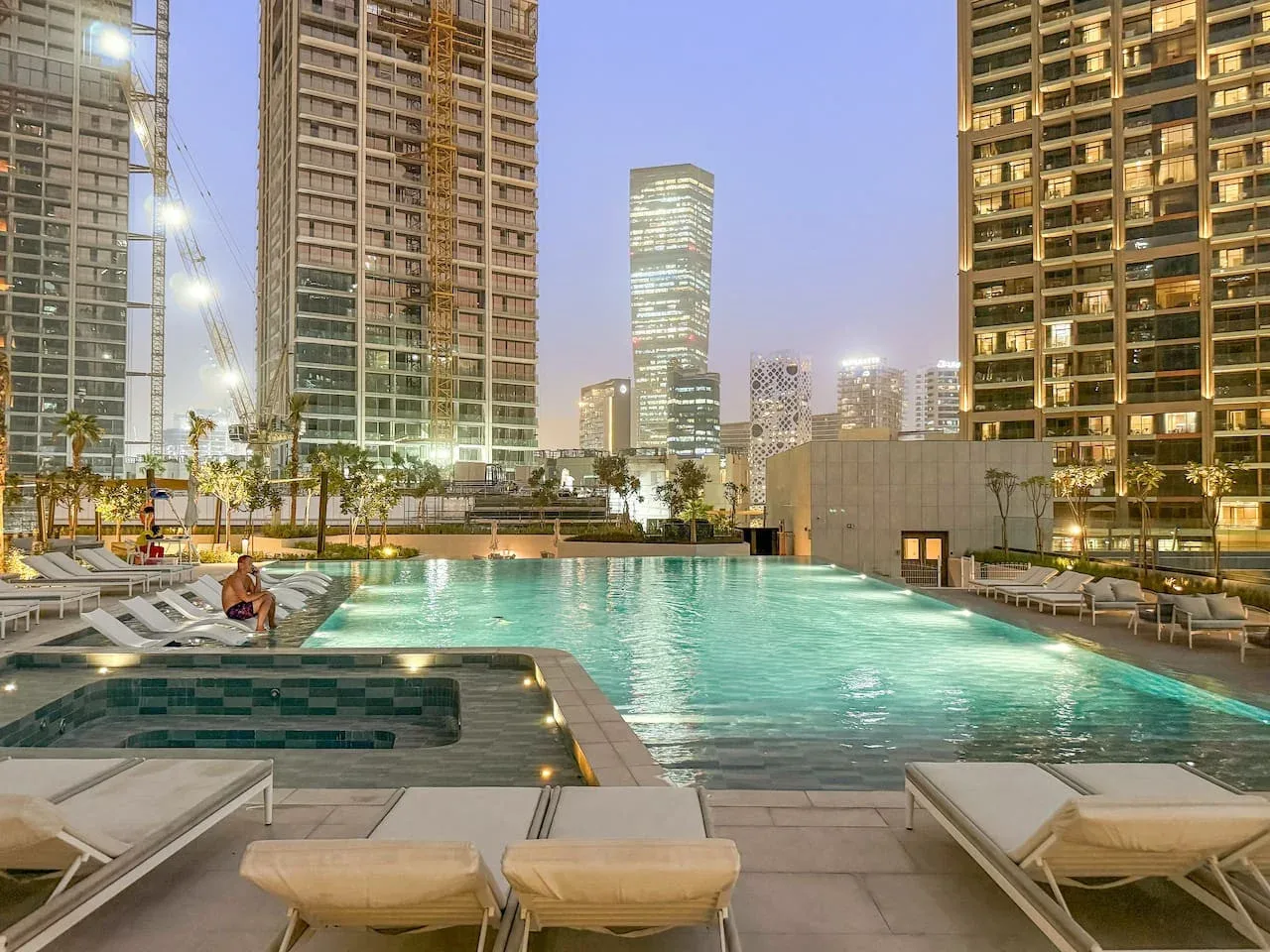 A rooftop swimming pool is surrounded by lounge seating and illuminated high-rise buildings.