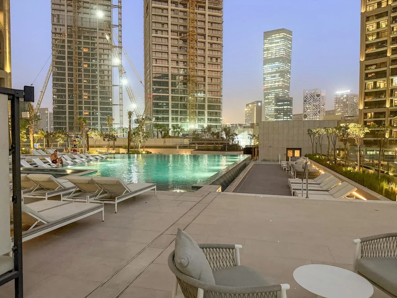 A rooftop pool deck with lounge seating overlooks illuminated city towers at dusk.