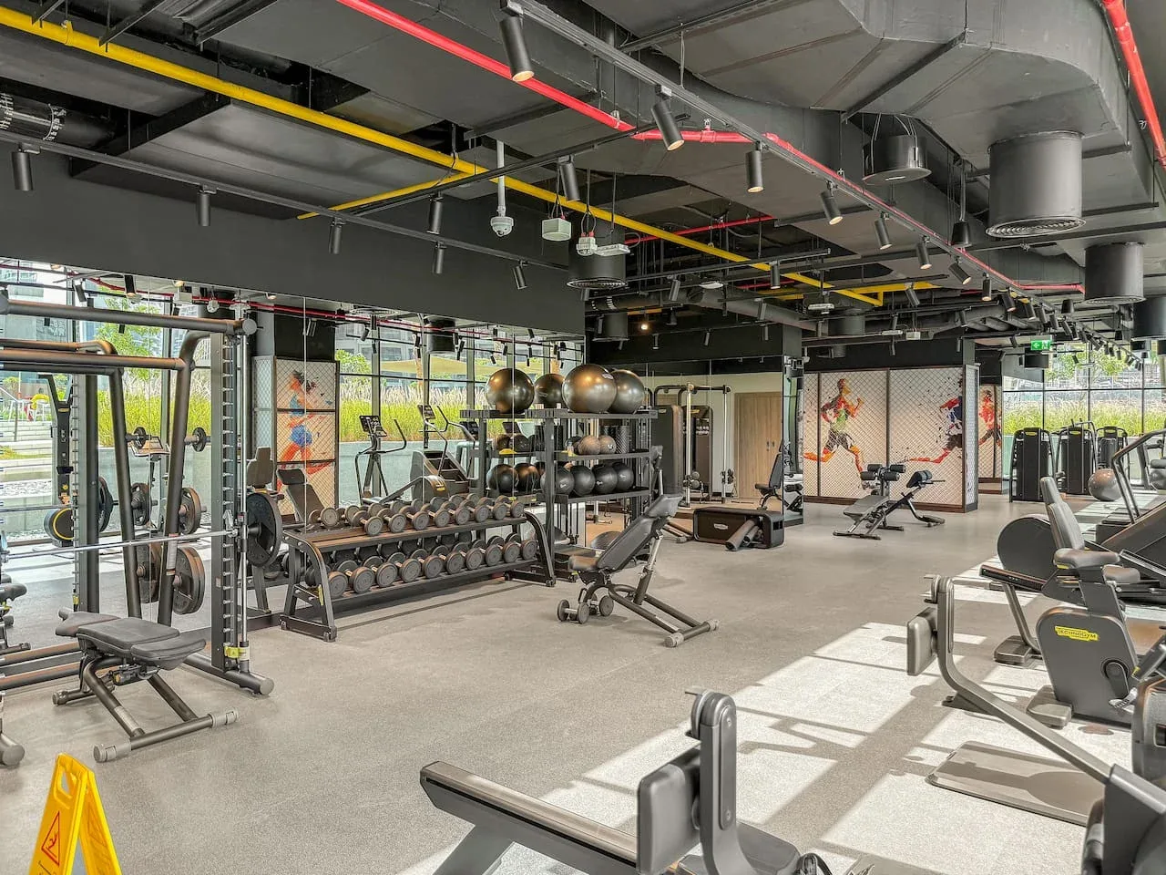A gym floor with weight racks and exercise machines beside large windows.