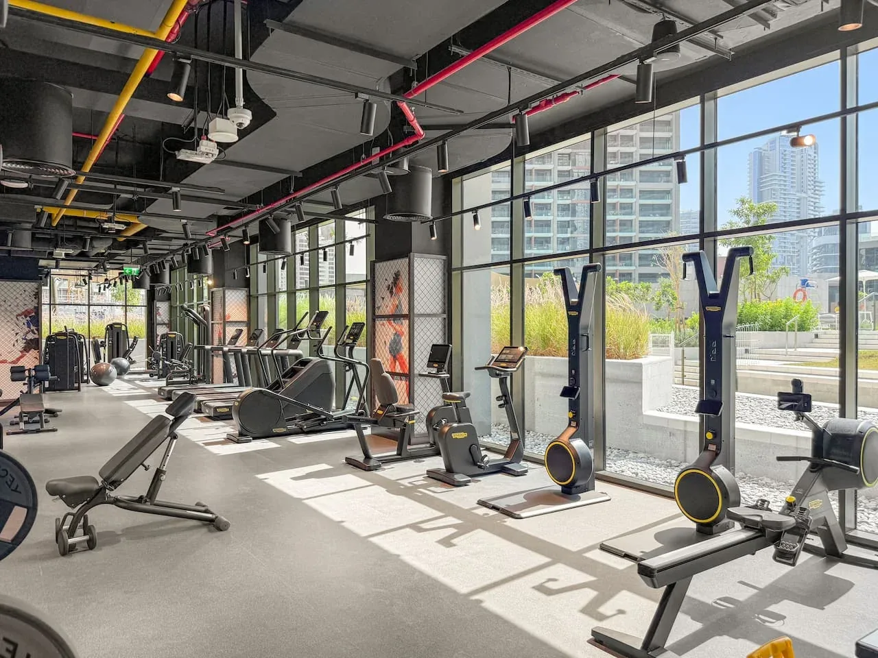 A bright gym with exercise machines beside large windows overlooking the city.