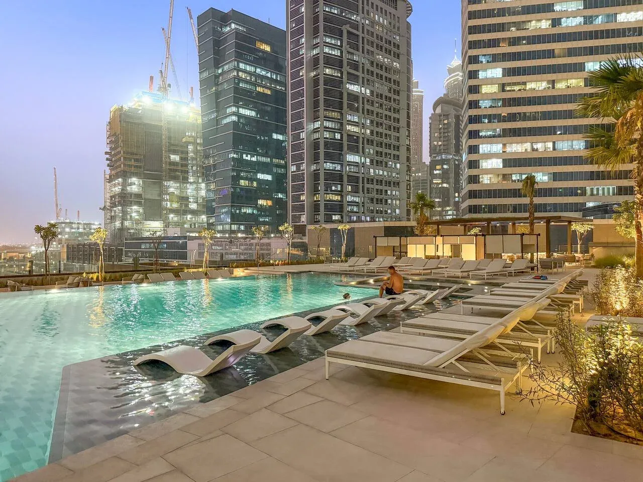A rooftop pool with lounge seating overlooks illuminated high-rise buildings.