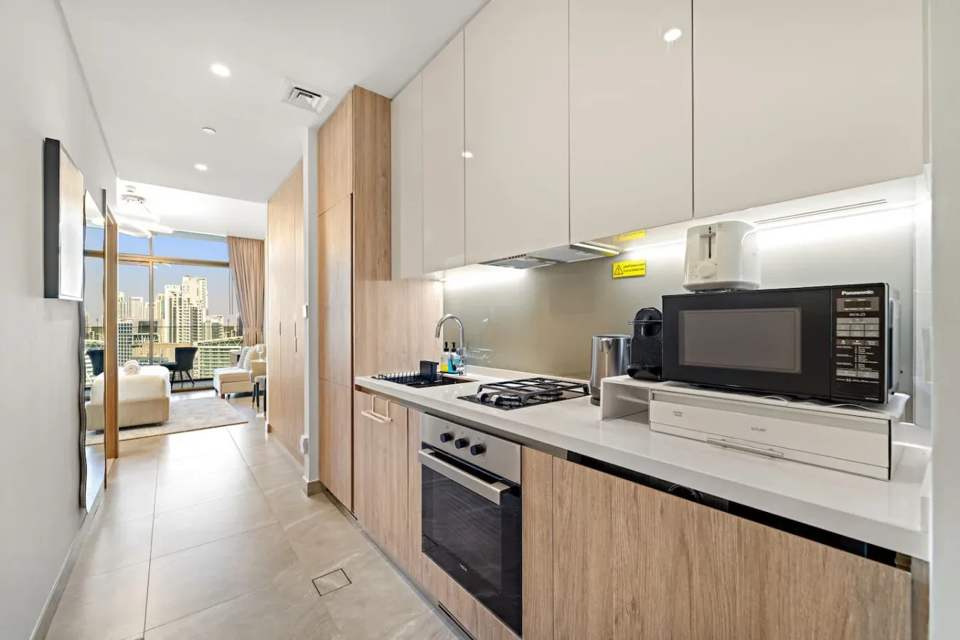 A modern kitchen with light cabinetry opens onto a living area with large windows.