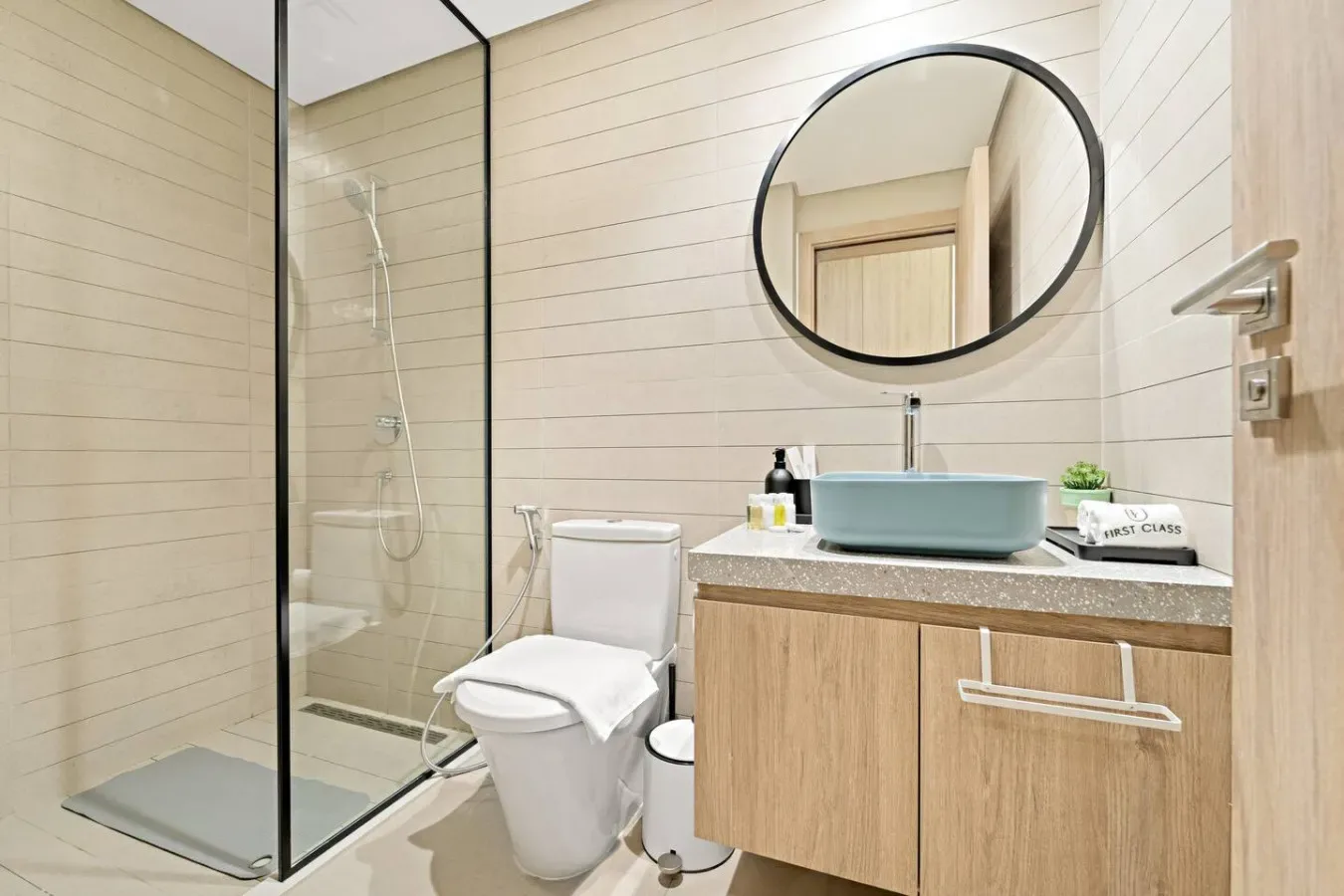 A bathroom with a glass shower, toilet, and wood vanity beneath a round mirror.