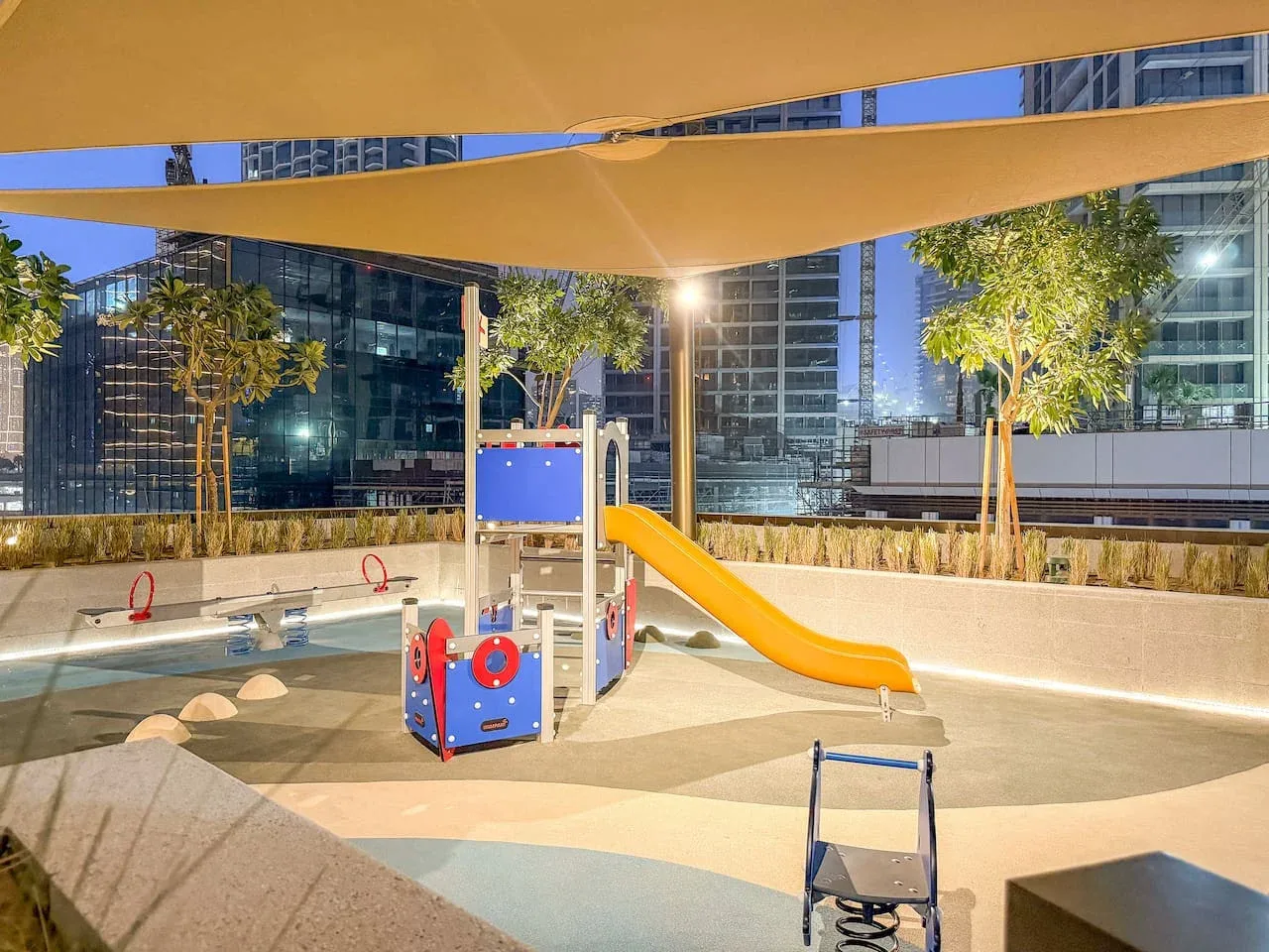An outdoor play area with a colorful play structure and slide beneath shade sails.