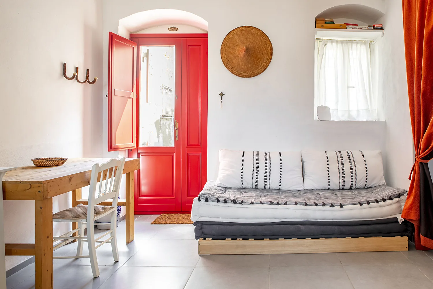 Una habitación luminosa con una cama baja, una mesa de madera y una puerta roja abierta.