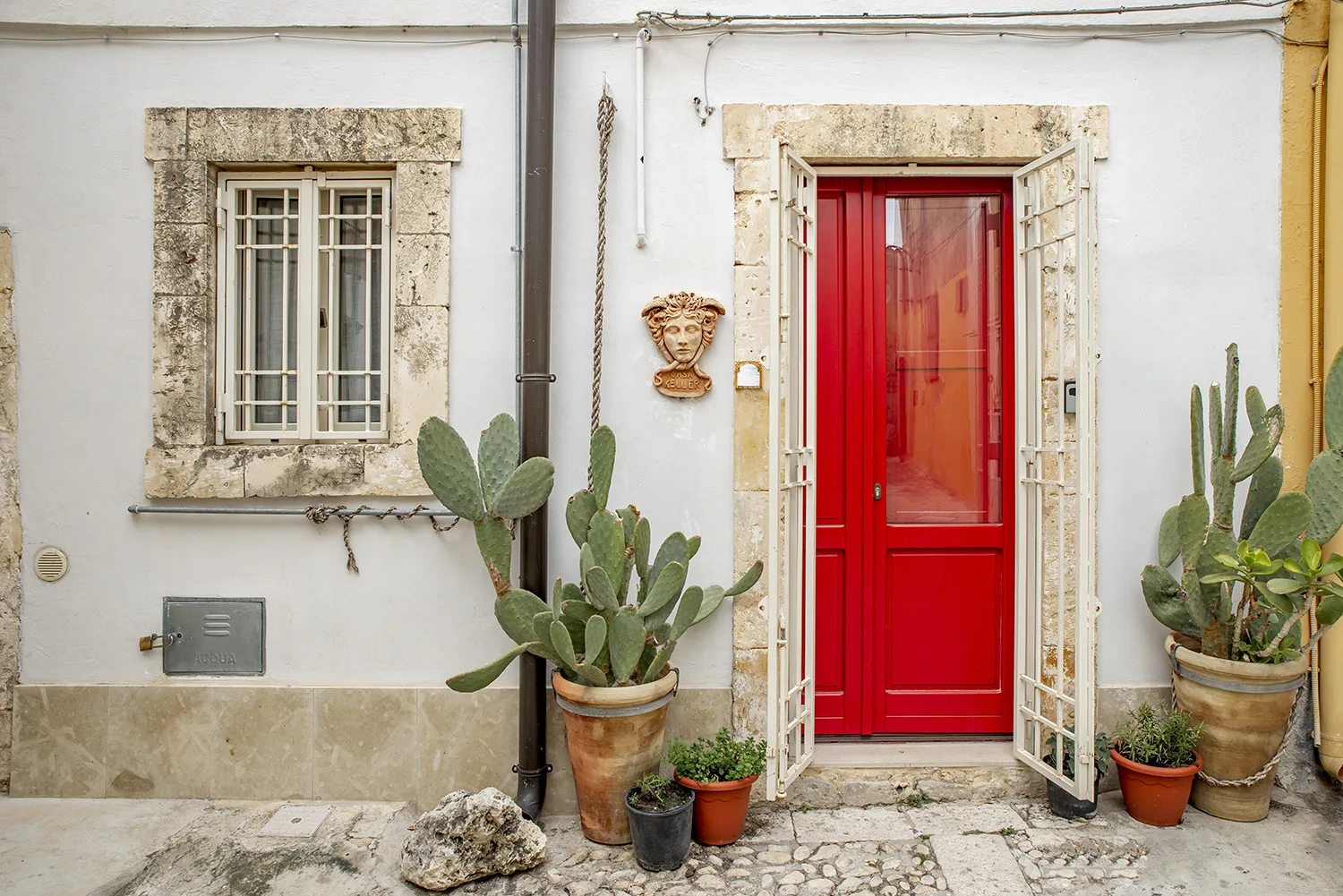 A white exterior wall with a red entrance door, barred window, and potted plants.