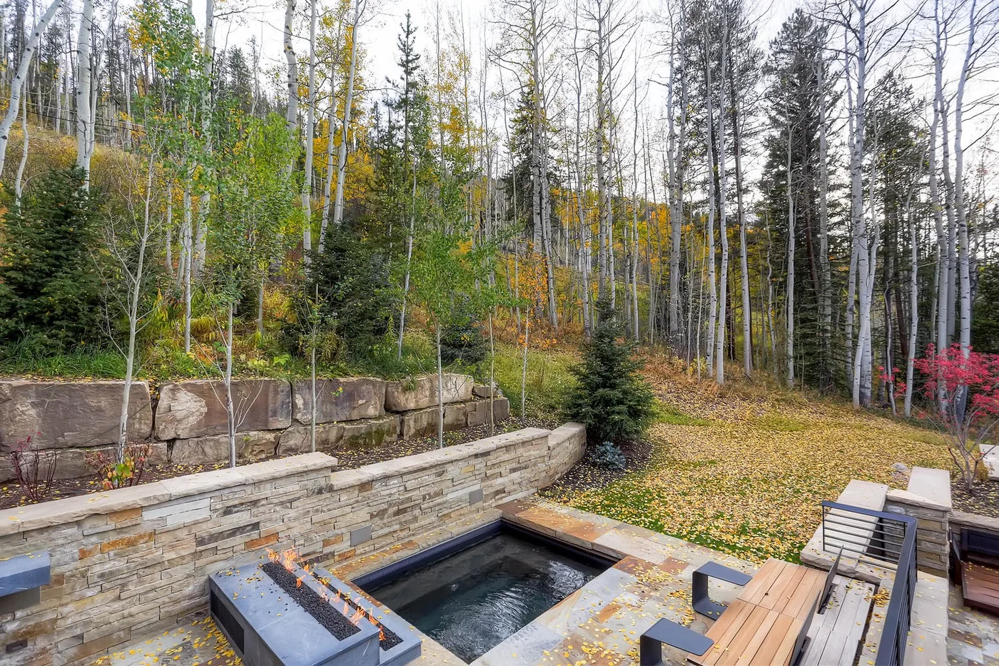 Una bañera de hidromasaje exterior está junto a un muro de piedra en un entorno arbolado.