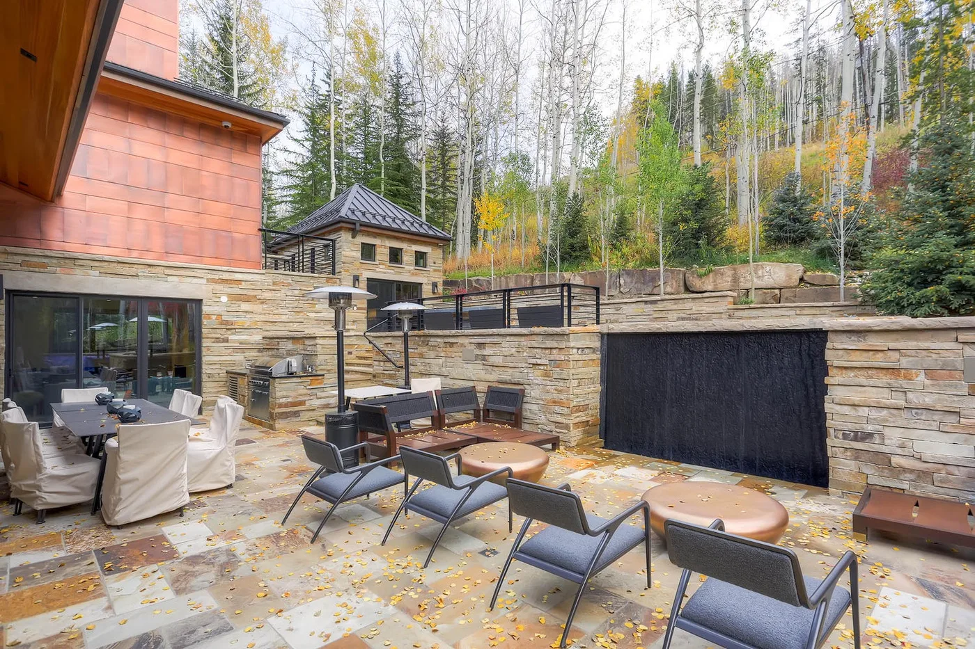 Un patio comedor al aire libre con mesas y asientos junto a un muro de piedra.