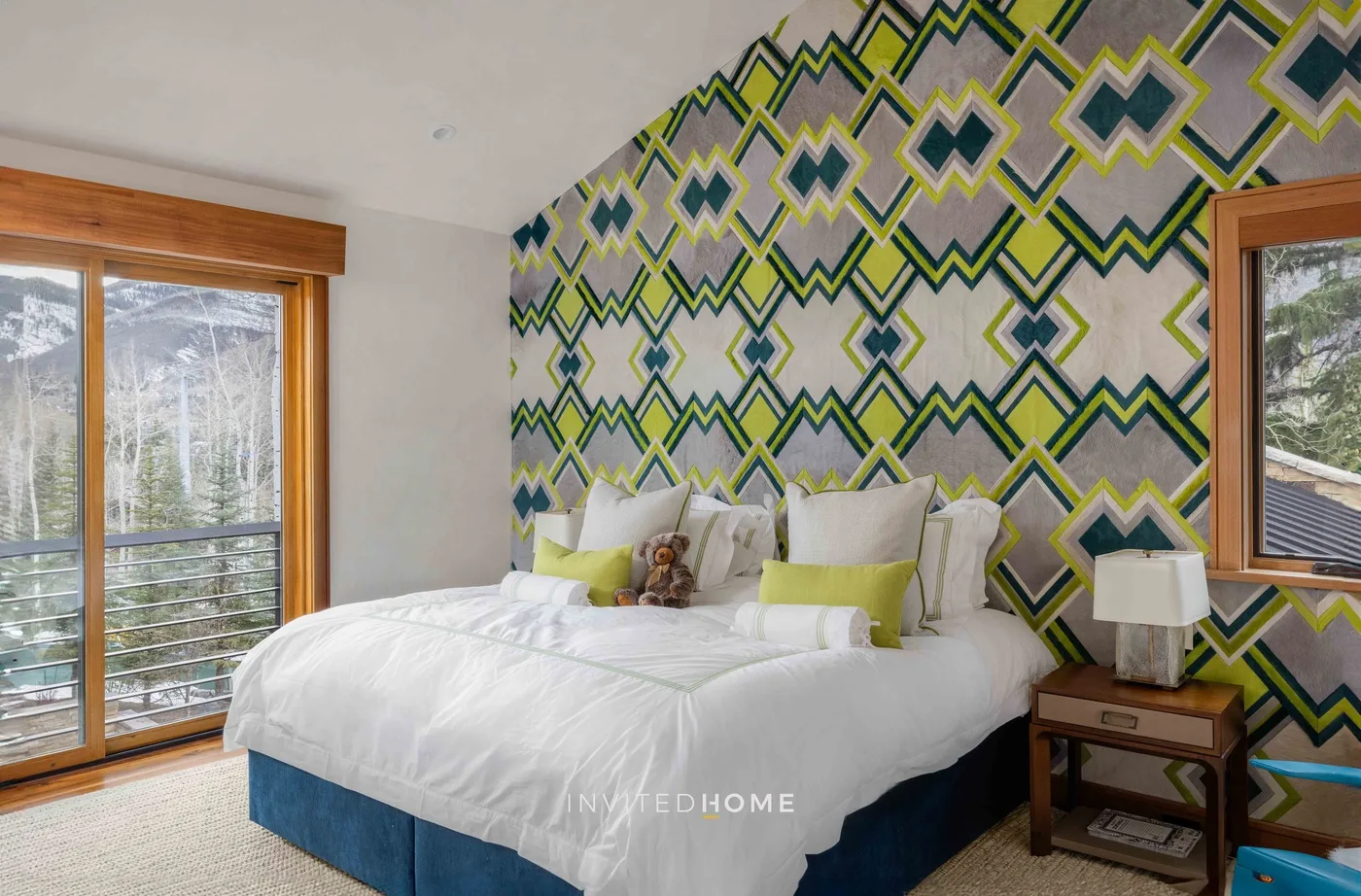 Un dormitorio con una cama blanca, una pared decorativa estampada y ventanas con vista a los árboles.