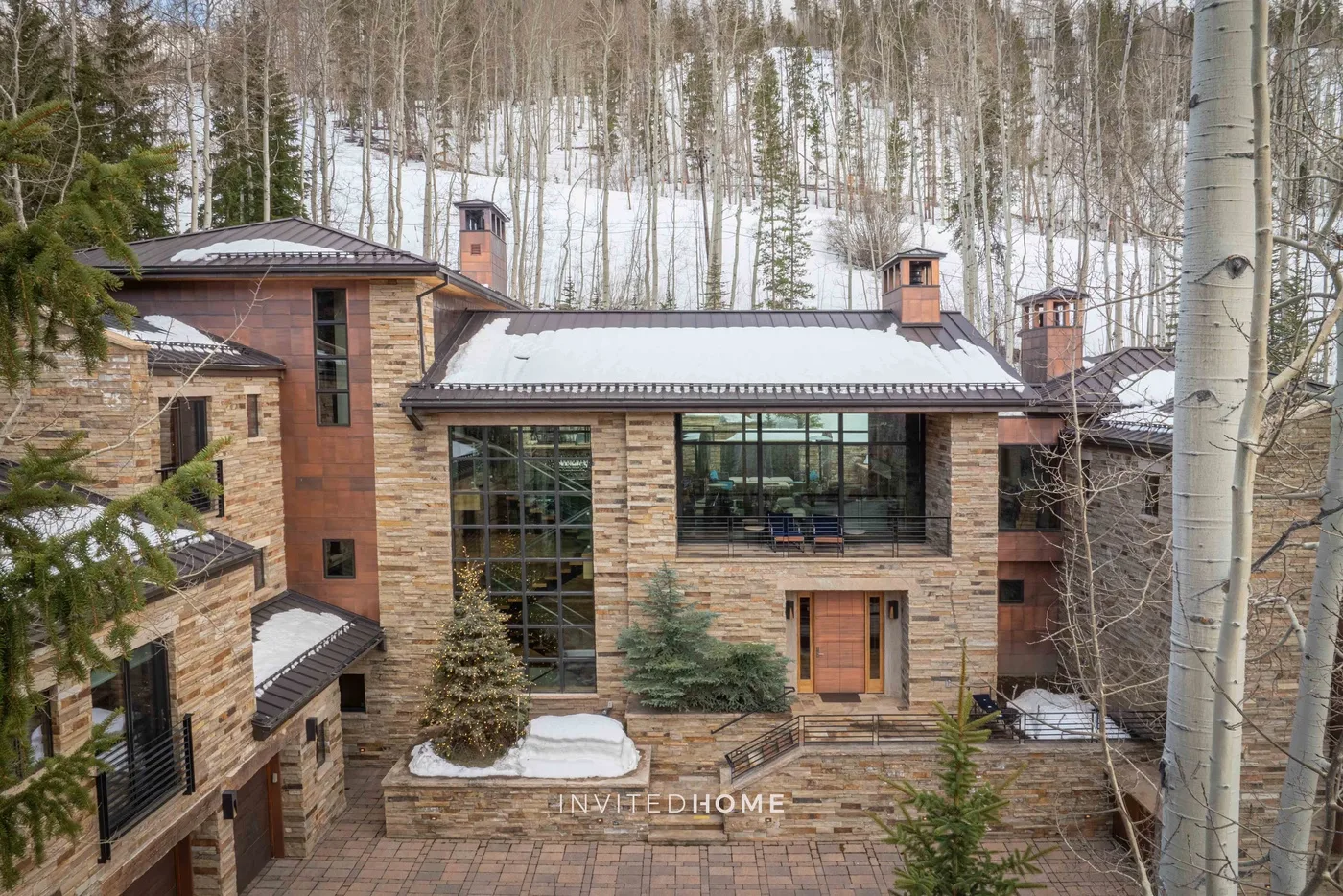 Una casa revestida de piedra da a un patio pavimentado, con un bosque nevado detrás.