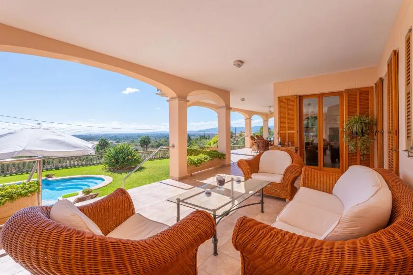 A covered terrace with wicker seating overlooks a swimming pool and garden.