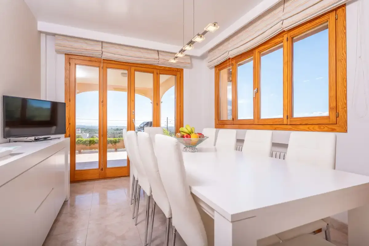 A bright dining area with a long white table beside large windows.