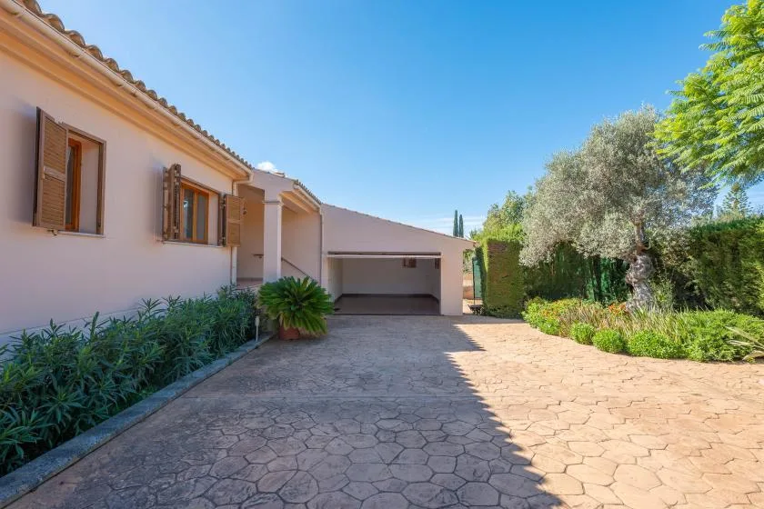 A paved driveway leads to a house with a covered parking area and garden greenery.
