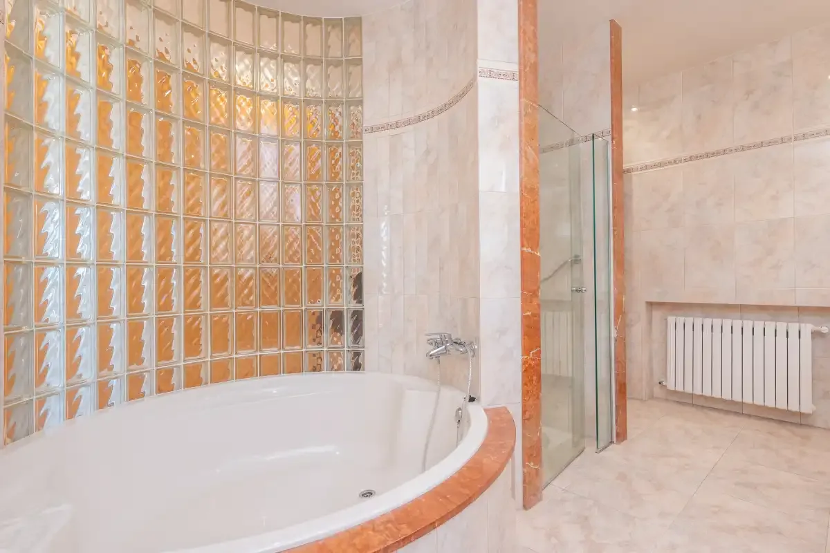 A bathroom features a curved bathtub, glass-block wall, and radiator.