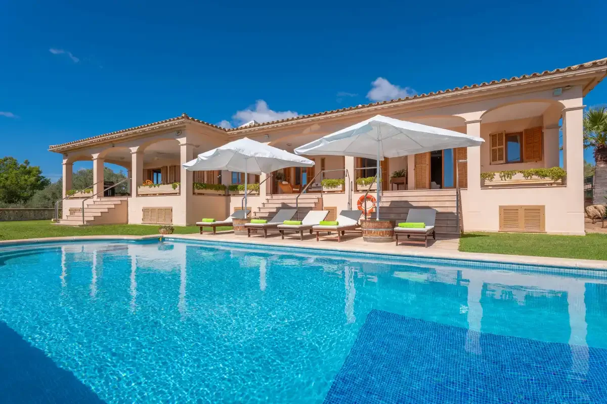 A swimming pool in front of a villa, with loungers and umbrellas beside it.
