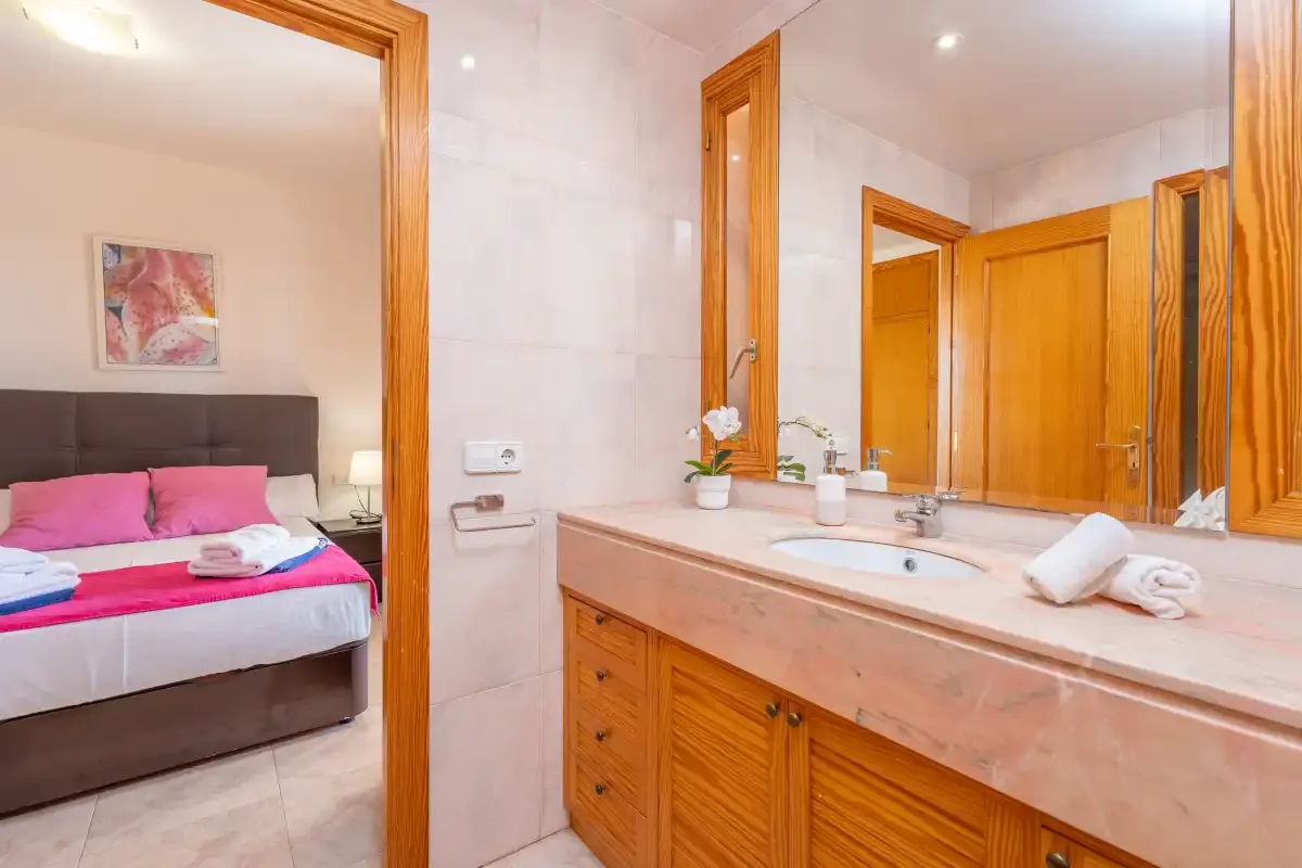 A bathroom vanity with a wide mirror overlooks a bedroom visible through the doorway.