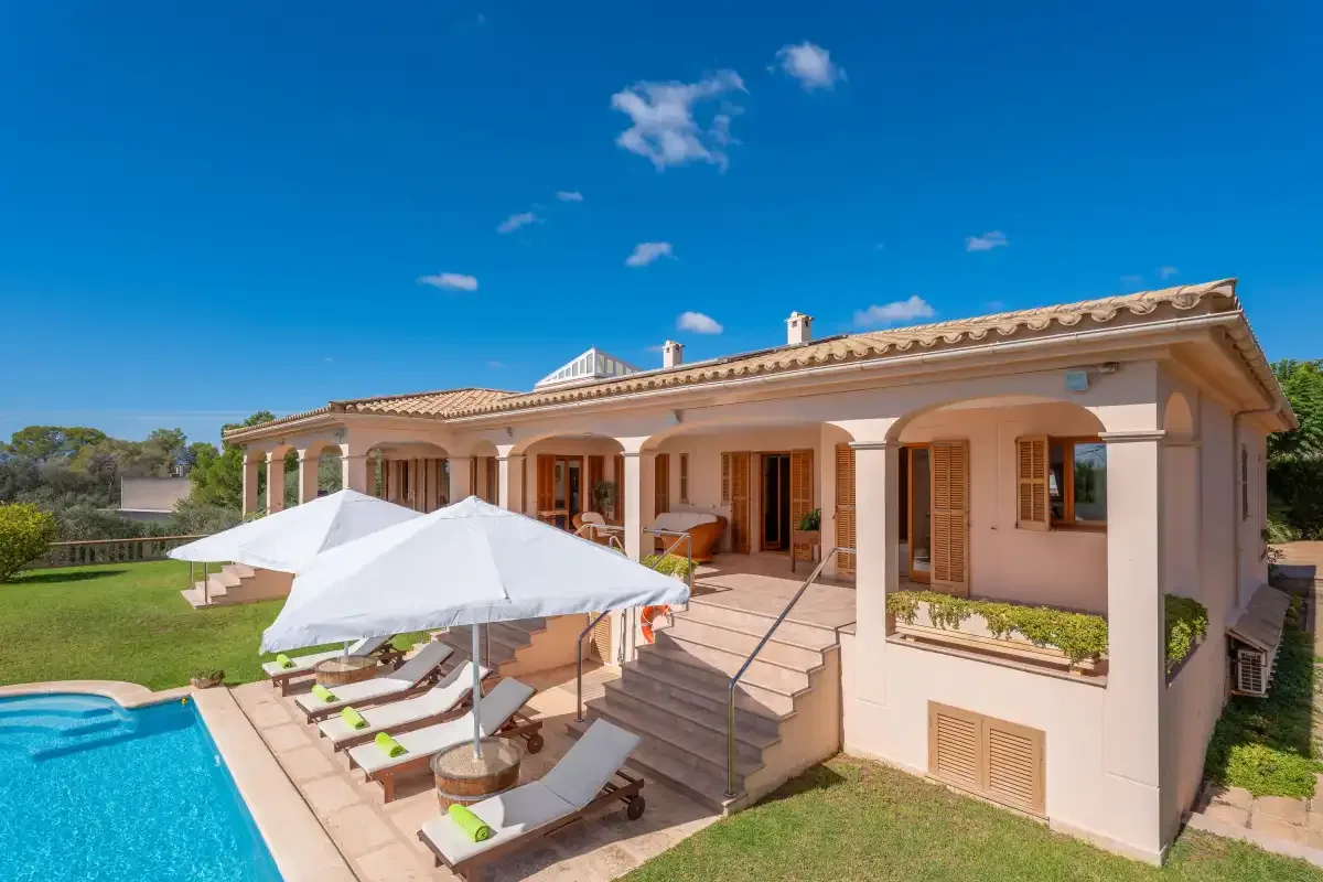 A pink house opens onto a patio with poolside loungers beneath a large white umbrella.