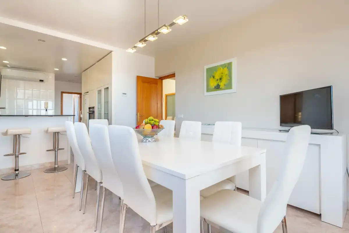 A bright dining area with white seating, a table, and an adjoining kitchen.