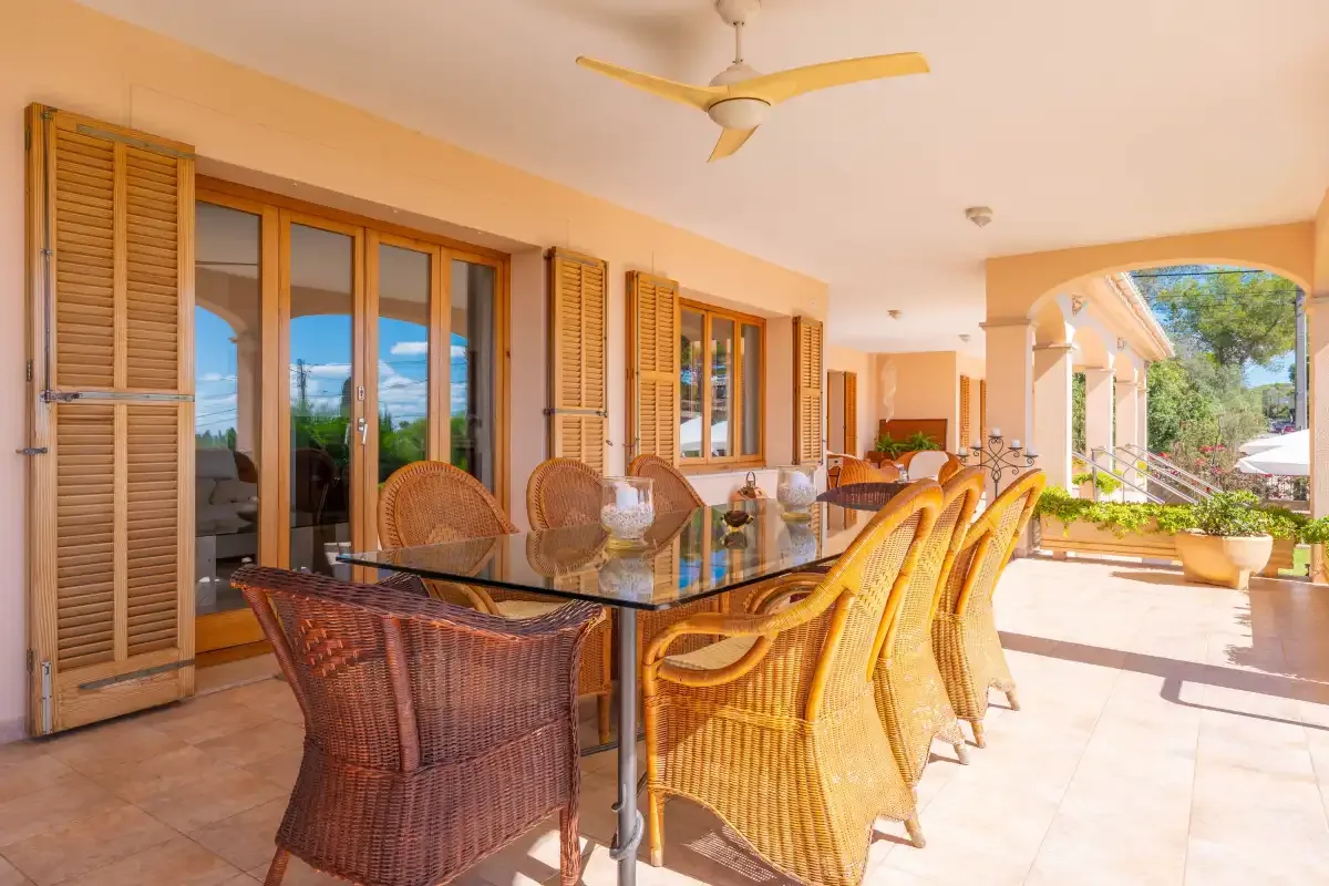 A covered outdoor dining area has a glass table and wicker seating.