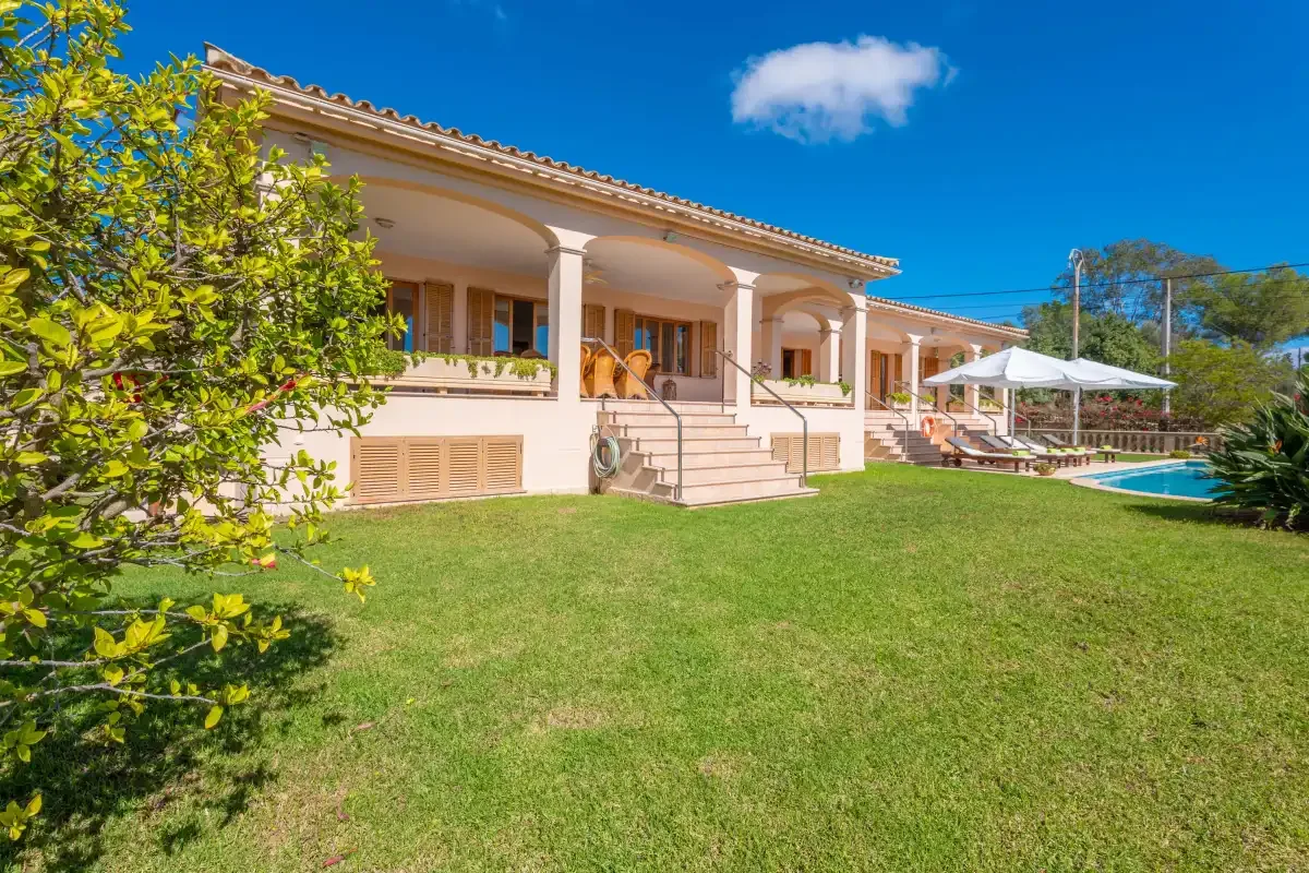 A villa with a covered terrace overlooks a lawn and swimming pool.