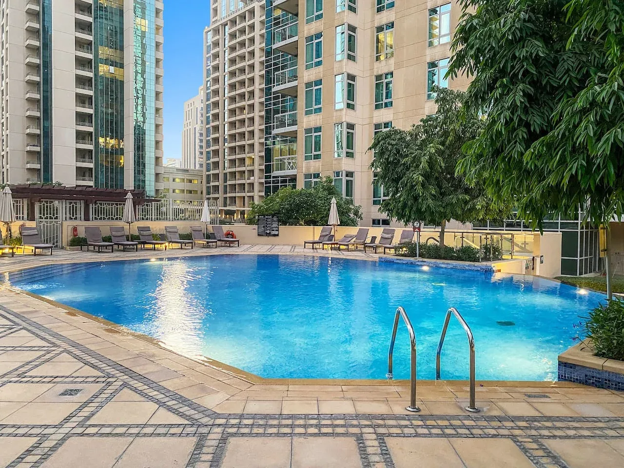 An outdoor swimming pool sits among high-rise buildings, with lounge seating along the deck.