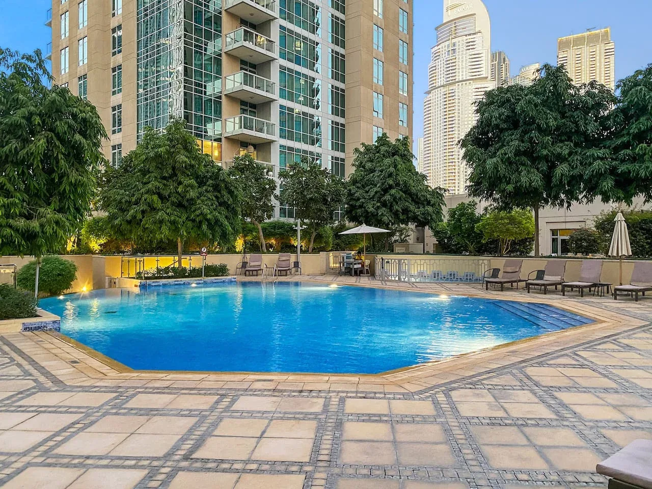 An outdoor swimming pool is surrounded by a paved deck, seating, and high-rise buildings.