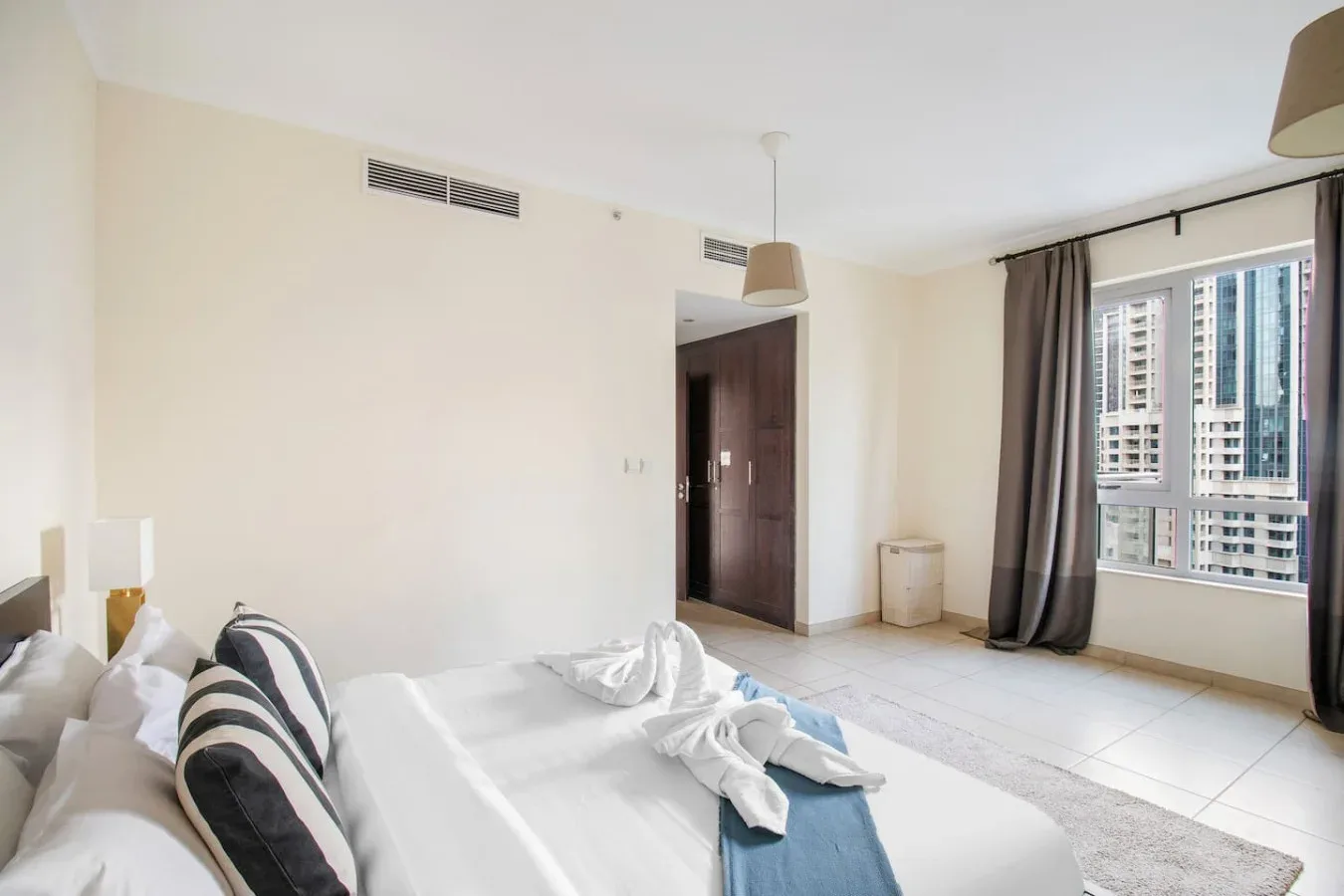 A bedroom with white bedding and a large window overlooking nearby buildings.