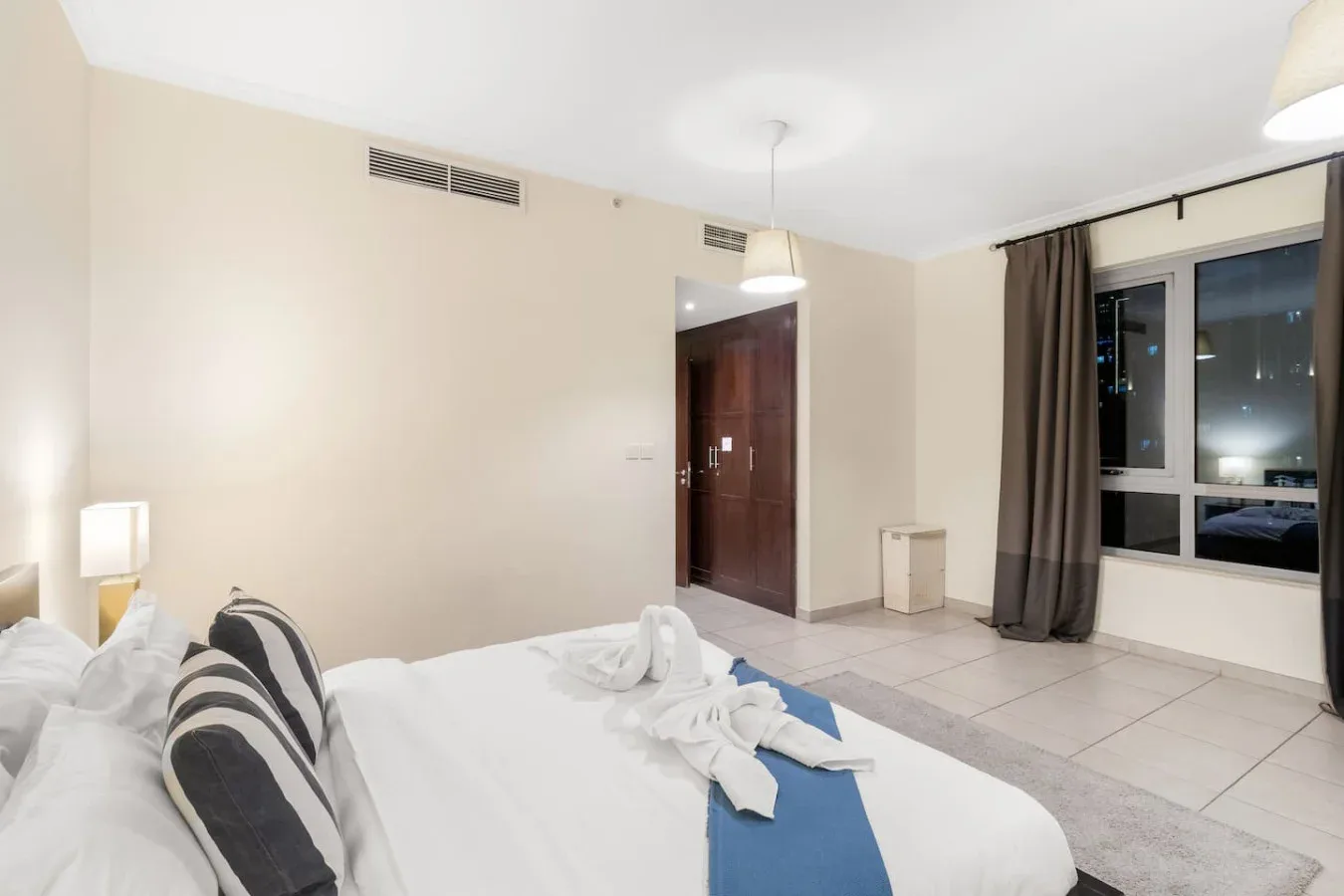 A bedroom with a neatly made bed and a large window overlooking city lights.