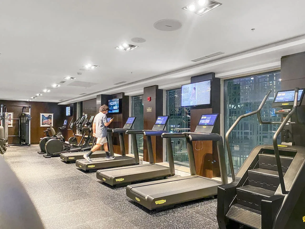 A gym with treadmills and other exercise equipment beside large windows.